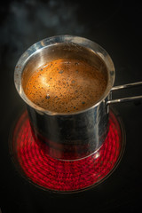 Stainless steel cezve with coffee in process of making standing at hot and red electrical stove in darkness, closeup, details