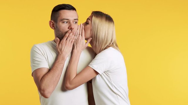 Young woman whispering secrets to surprised man over yellow background