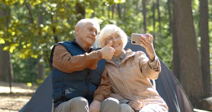 Cheerful old Caucasian married couple taking selfie photo on smartphone at camping in forest. Senior male and female tourists sitting in camp at tent in wood and making picture on phone camera.
