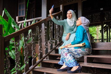 Muslim Malay women wearing hijab and traditional costume are taking selfie during Aidilfitri celebrations at the terrace of traditional wooden house.