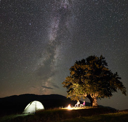 Romantic couple hikers having a rest at summer night camping in the mountains. Man and woman sitting on chairs beside bonfire, tourist tent under big tree and starry sky full of stars and Milky way.