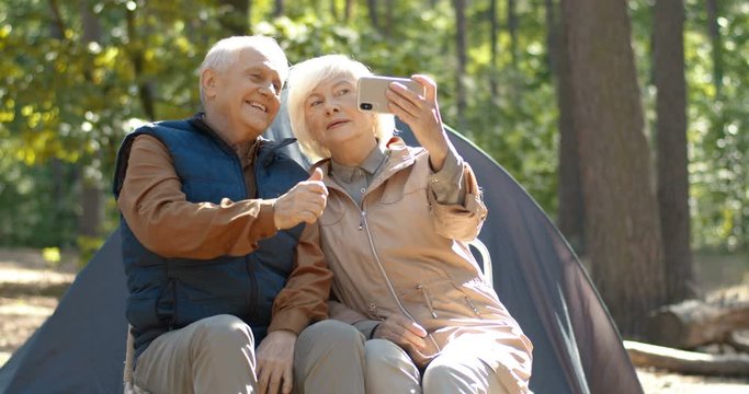 Elderly male and female Caucasian campers resting at tent and making selfie on smartfone. Happy senior couple having rest in wood and fooling around while taking photo on phone camera. Camping concept