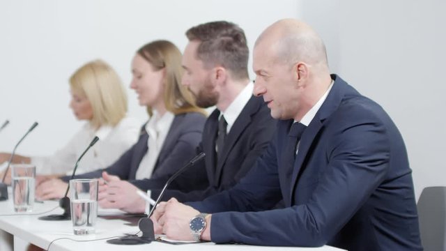 Rack focused shot of experienced businessman in formal suit talking into microphone while sitting at desk with group of colleagues during press conference