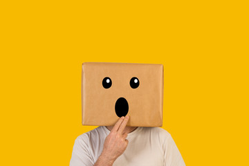 Hombre con caja de cartón en la cabeza con una expresión de asombro en la cara sobre fondo amarillo liso brillante. Vista de frente. Copy space - obrazy, fototapety, plakaty