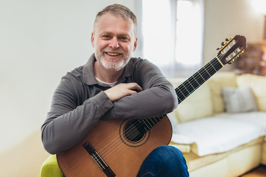 Handsome mature man in casual clothes is smiling while playing guitar at home