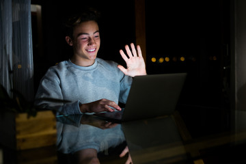 one caucasian man or teenager waving at someone doing videocall with someone in quarantine and lockdown - studying and doing homeowrk or working at night at home in videocall with his class online - obrazy, fototapety, plakaty