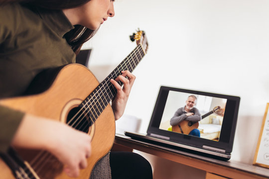 Focused girl playing acoustic guitar and watching online course on laptop while practicing at home. Online training, online classes.