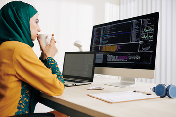 Pensive female muslim coder drinking tea and looking at programming code on computer screen
