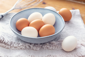 Close up of six white and brown eggs in a light blue bown placed on a dish-cloth with a metal mixer in the background
