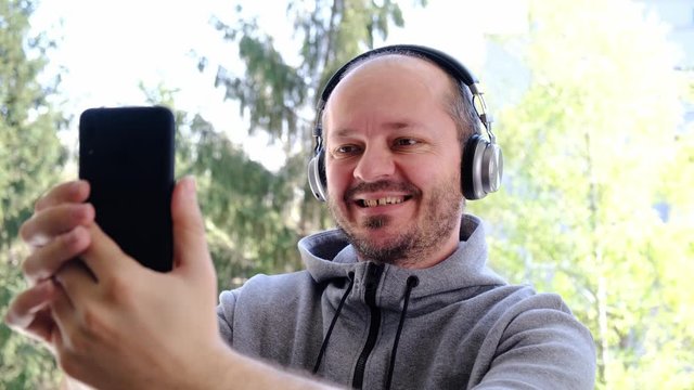 Bearded adult man in hoodie with wireless headphones taking selfie with smart phone in park outdoors, head and shoulders