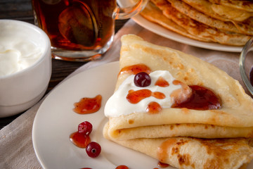 several pancakes with berries, berries, cranberries,  sour cream and jam on a plate,  mug of black tea  on a dark wooden