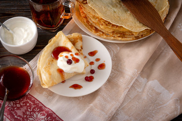 lot of pancakes with berries, cranberries, cranberries, sour cream and jam on a plate, a mug of black tea on a towel on a dark wooden background