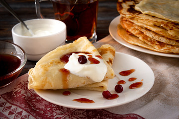 lot of pancakes with berries, cranberries,sour cream and jam on a plate, a mug of black tea on a towel on a dark wooden
