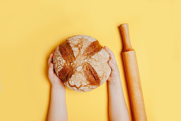 Tasty big round bread in child's hands on yellow background. Top view, flat lay