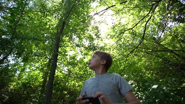 Young white kid taking photos of beautiful green landscapes standing alone in old green forest. Boy using digital camera of smartphone.