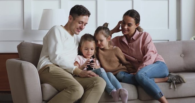 Happy young married couple relaxing on couch with adorable small children siblings, watching funny cartoons on smartphone. Smiling young father recording video, taking selfie with cute kids and wife.