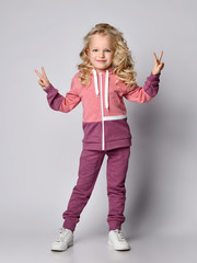 Curly hair blonde kid girl in modern fashion pink gray sports suit shows V-sign victory gesture with both hands