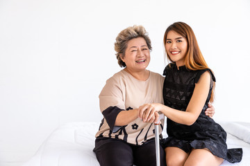 Happy senior elderly Asian woman female mother holding cane with daughter sitting on bed in bedroom, looking at camera