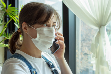 Portrait of young woman in a mask who is talking on the phone while standing by the window,...