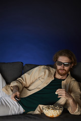 Obraz premium Vertical portrait of modern bearded man watching movie and wearing stereo glasses while lying on couch in dark room, copy space