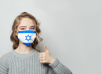 Happy woman in face mask holding thumb up with national flag Israel background. Flu epidemic and virus protection concept