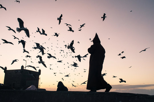 Silueta de un hombre misterioso encapuchado con ropa tradicional al atardecer y gaviotas volando