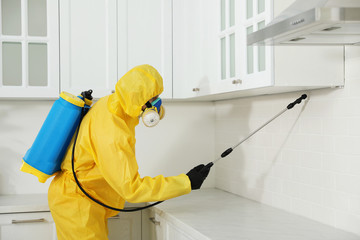 Pest control worker in protective suit spraying insecticide on furniture indoors