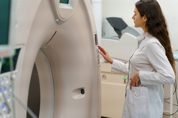 Medical resident does computer tomography fot patient. Young girl in white coat looks in CT and press button to start procedure research diagnostic