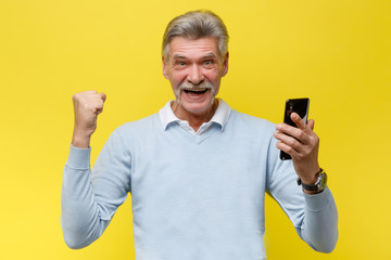 Emotional senior man with phone winn something while posing on yellow background