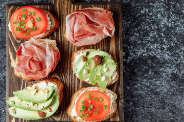 Assorted Italian bruschette snack on a wooden chopping board on a dark textured table. Mini sandwich mix. Close up