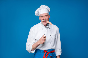 Portrait of a smiling senior chef holding sausage on special fork, isolated on blue background