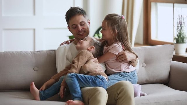 Smiling young daddy holding on lap, embracing little preschool kids daughters, enjoying sweet tender moment together in living room. Happy father relaxing on sofa with cute small children girls.