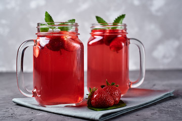 Homemade strawberry tea with mint in mason jar on gray concrete table background, copy space. Cold summer berry drink in sparkling glasses. Fresh vitamin beverage