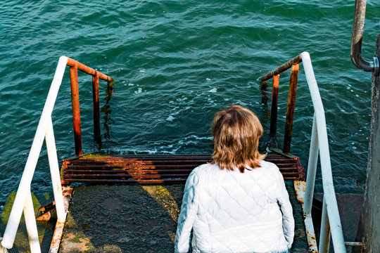 Woman sitting on the rusty staircase into the water in sea. Back view. Dreaming. Thinking. Meditation. Peaceful place. Focus on the water and stairs. Path. Ladder with steps. Waves. Jacket - Powered by Adobe