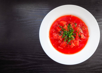 Round white plate with red borscht and chopped green onions on a dark background. National cuisine