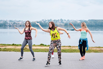 A group of young women, wearing colorful sports outfits, doing zumba exercises outside by city lake. Dancing training to loose weight in summer. Healthy lifestyle concept. Female sport leisure.