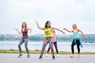 A group of young women, wearing colorful sports outfits, doing zumba exercises outside by city lake. Dancing training to loose weight in summer. Healthy lifestyle concept. Female sport leisure.