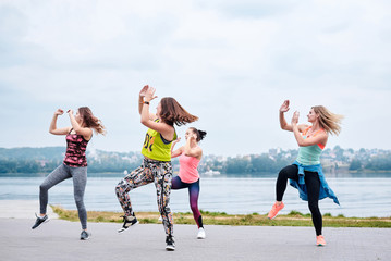 A group of young women, wearing colorful sports outfits, doing zumba exercises outside by city lake. Dancing training to loose weight in summer. Healthy lifestyle concept. Female sport leisure.