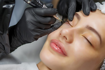 Permanent make up eyeliner procedure, applying on young girl. Close-up. Young beautiful woman making permanent makeup in cosmetology salon. Young girl applying permanent eyeliner in beauty studio