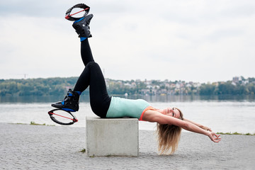 Young blond fit slim woman in kangoo jumps, sitting by city lake in summer. Fitness trainer,...