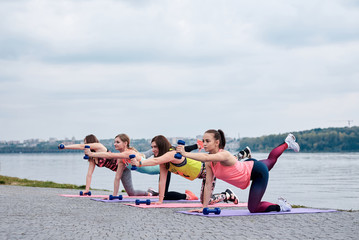 A group of four young women, wearing colorful sports outfit,doing fitness exercises on yoga mats outside by city lake in summer. Workout power female training to loose weight and body shape at nature.