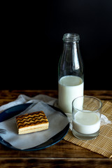 on a black background on a wooden table is a glass bottle with milk, next to a glass of milk and a cake