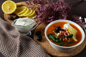 Soup Solyanka is a combined meat soup with smoked meat , various types of meat and tomato paste, serving dishes with sour cream, lemon and fresh herbs. Black olives on the table and a sprig of fresh f