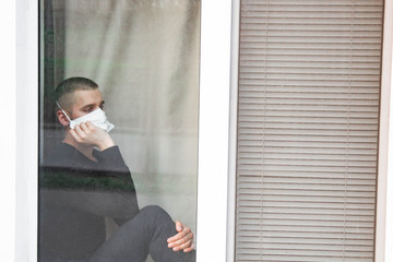 Upset young man sitting by the window and looking away. The concept of loneliness in quarantine during the pandemic of the coronavirus COVID-19.