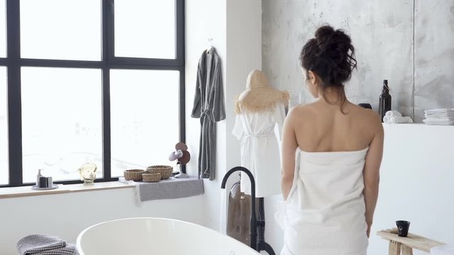 Woman dressed in the white shower towel walking to the buttub. View from back. Handheld shot.
