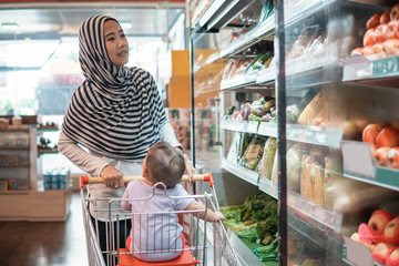 happy infant baby sitting in shopping cart or trolley in grocery supermarket with mother