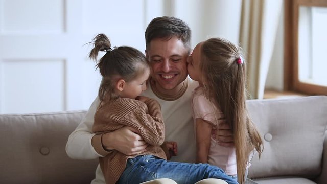 Happy young handsome father sitting on sofa, holding on lap small children daughters, enjoying tender sweet moment at home. Cute little kids girls embracing kissing smiling father, slow motion.