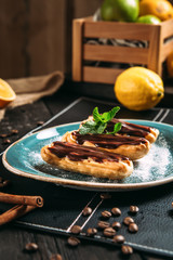 Side view of yummy eclairs cakes with chocolate on a blue plate on a black napkin background, vertical