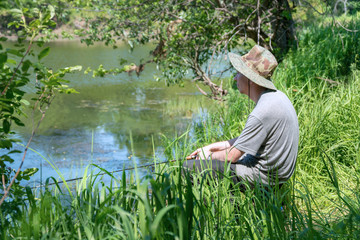 Naklejka na ściany i meble A fisherman in a hat is fishing on a forest lake or river