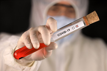 The doctor in a protective suit and mask holding a glass tube with a positive blood test result for...